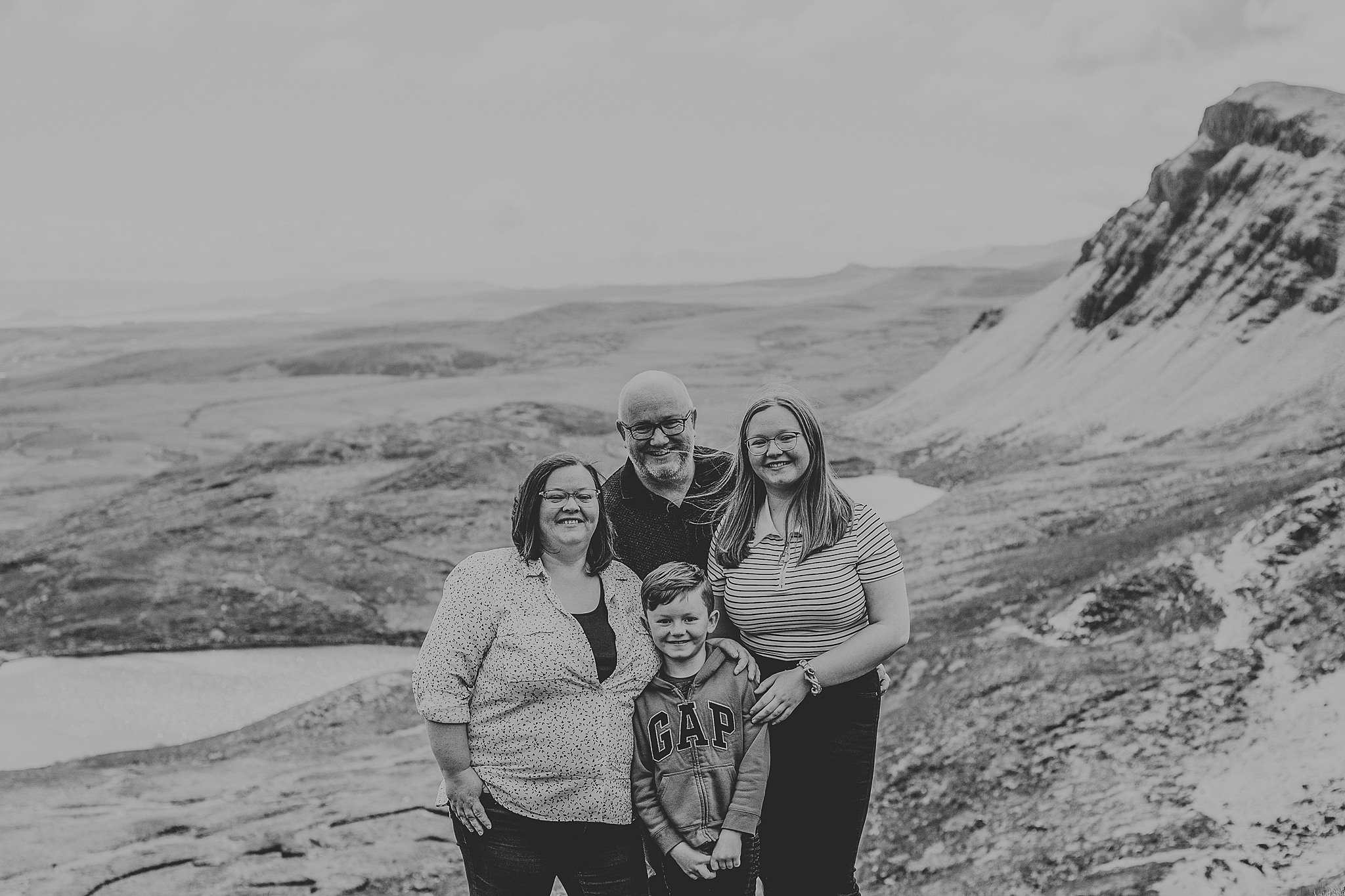 black and white photo of a family of four smiling and hugging at the quiraing on the isle of skye