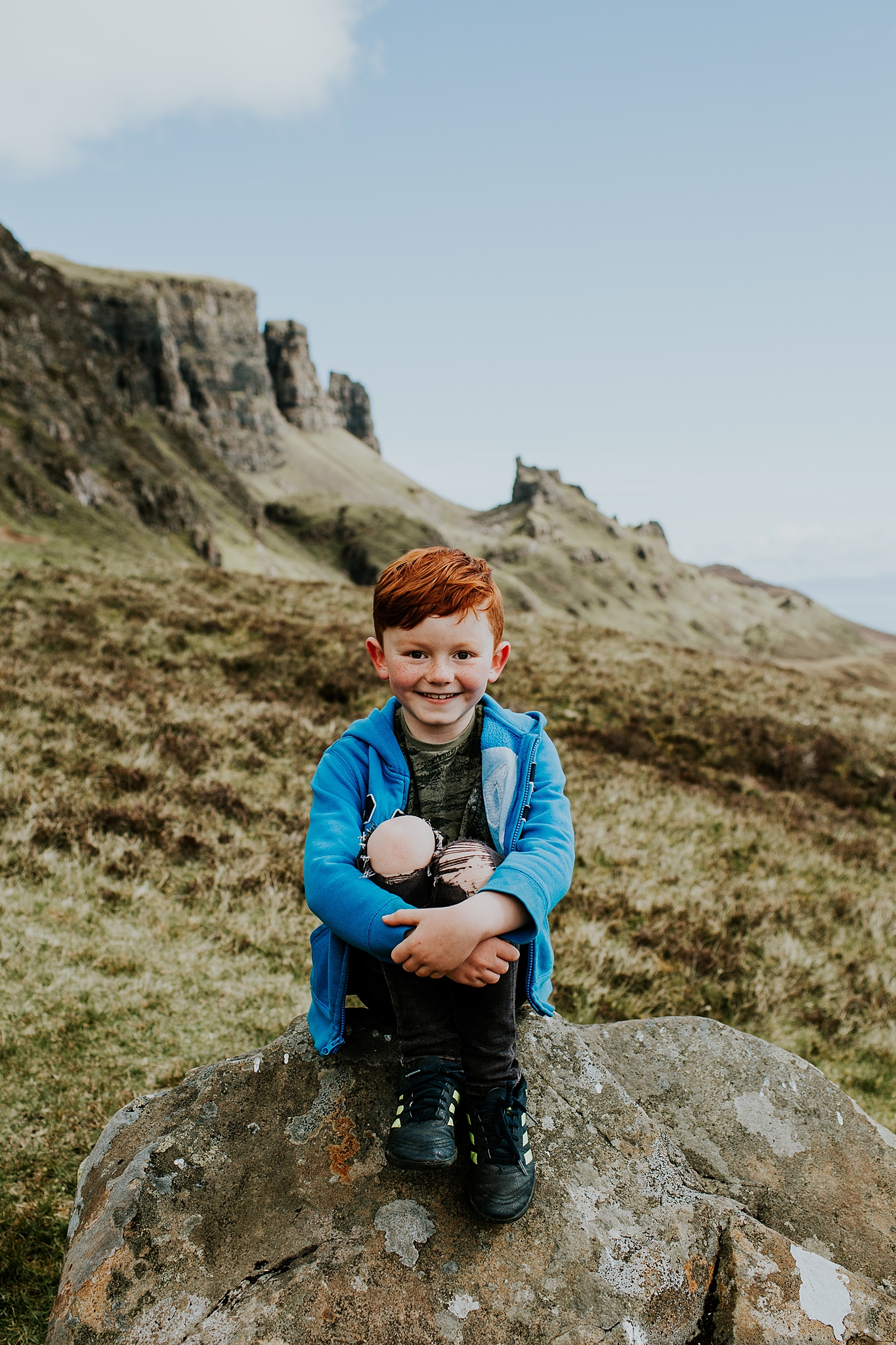 young red headed boy sitting on a rock smiling and holding his knees at the quiraing on the isle of skye