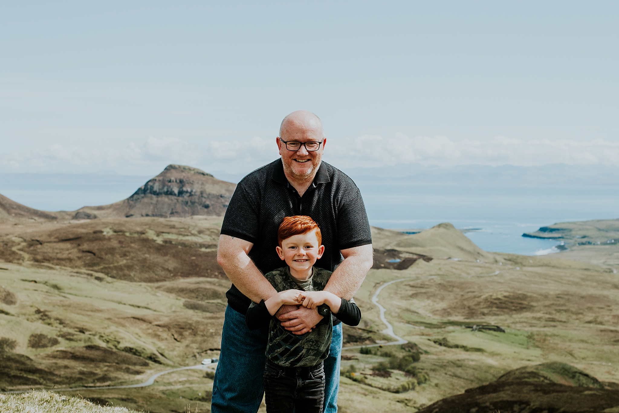 dad smiling with arms around his young red headed son also smiling in the sunshine at the quiraing on the isle of skye