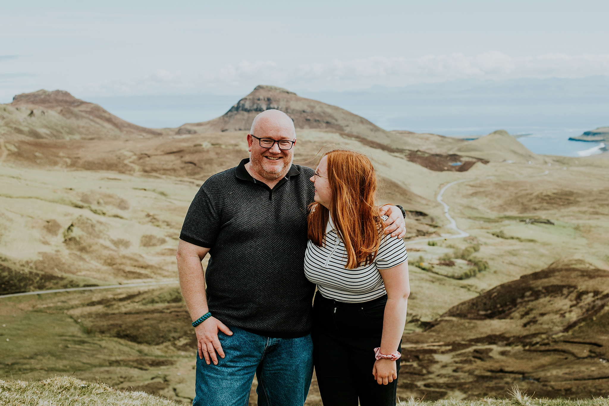dad smiling and hugging red haired daughter who is looking up at him overlooking staffin on the isle of skye from the quiraing